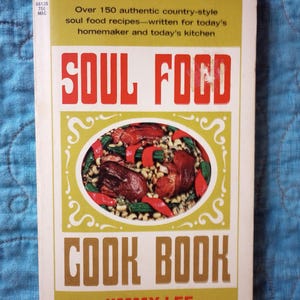 May include: A paperback cookbook titled Soul Food Cook Book by Jimmy Lee. The cover features a central oval illustration of a dish with meat, beans, and red peppers, surrounded by olive green and white borders with black and red text.