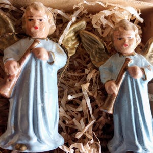 Puede incluir: Dos figuras de &aacute;ngeles vintage. Cada &aacute;ngel viste una t&uacute;nica azul claro y tiene alas doradas. Sostienen una trompeta marr&oacute;n. Los &aacute;ngeles est&aacute;n en una caja de cart&oacute;n con relleno de papel.