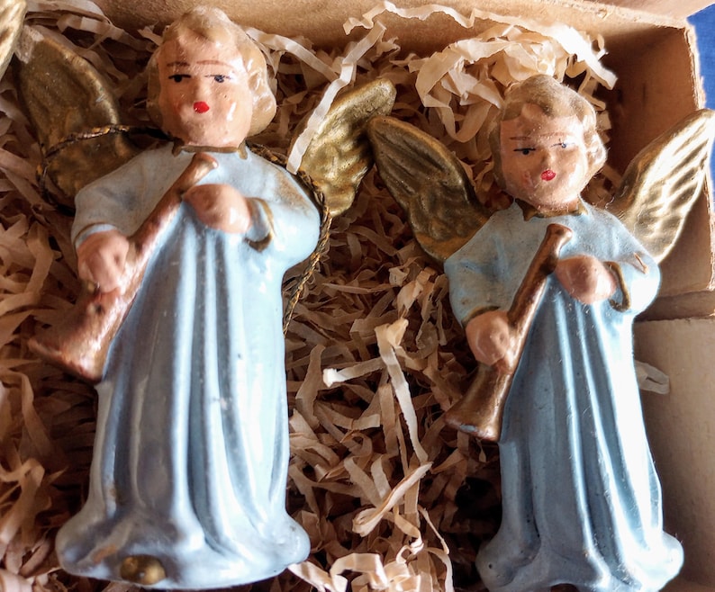 Puede incluir: Dos figuras de &aacute;ngeles vintage. Cada &aacute;ngel viste una t&uacute;nica azul claro y tiene alas doradas. Sostienen una trompeta marr&oacute;n. Los &aacute;ngeles est&aacute;n en una caja de cart&oacute;n con relleno de papel.