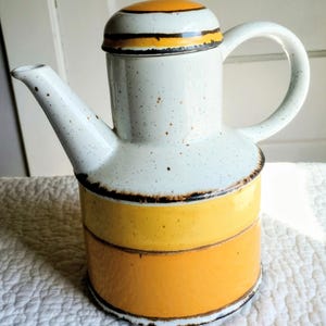 Könnte beinhalten: Eine Keramik-Teekanne mit zylindrischem Boden, horizontalen gelben und orangen Streifen und einem weißen Deckel. Der obere Teil ist weiß mit braunen Sprenkeln, einem geschwungenen Henkel und einem Ausguss, akzentuiert durch dunkelbraune Linien.