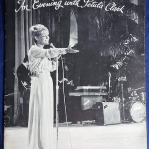 Peut inclure: Affiche de concert vintage en noir et blanc intitulée "An Evening with Petula Clark". L'image montre une femme en robe blanche sur scène, tenant un microphone. Des palmiers sont en arrière-plan.
