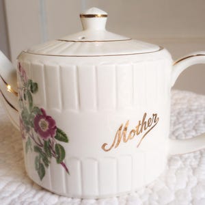 May include: Cream-colored ceramic teapot with a lid and handle. The teapot features a floral design with pink and green accents, and the word "Mother" in gold script. Gold trim accents the lid and handle.