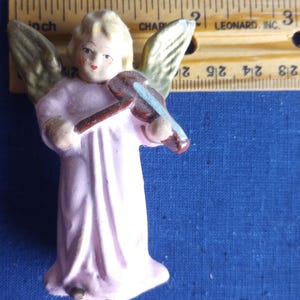 Puede incluir: Peque&ntilde;a figura de &aacute;ngel vintage con una t&uacute;nica rosa claro y alas doradas. El &aacute;ngel sostiene un viol&iacute;n marr&oacute;n con un arco azul. La figura mide aproximadamente 5 cm de altura.