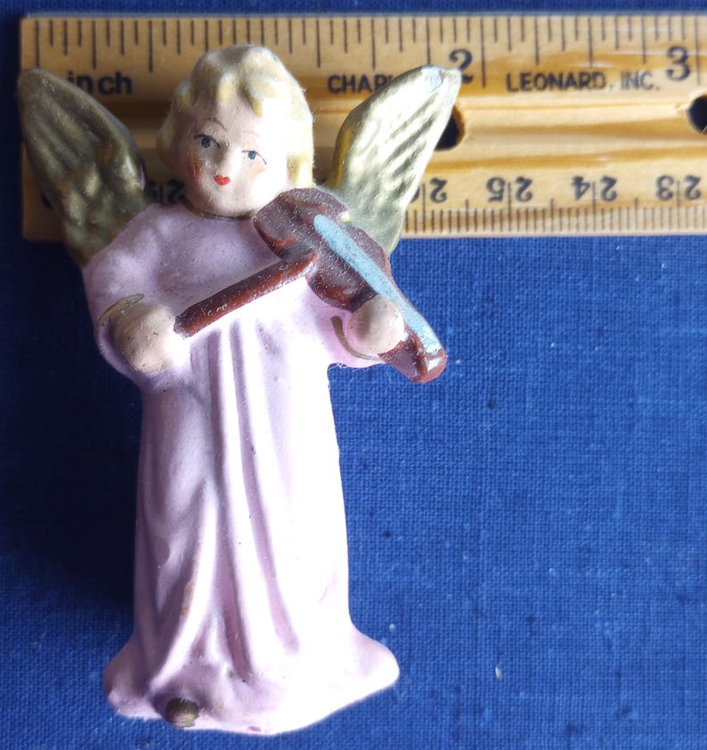 Puede incluir: Peque&ntilde;a figura de &aacute;ngel vintage con una t&uacute;nica rosa claro y alas doradas. El &aacute;ngel sostiene un viol&iacute;n marr&oacute;n con un arco azul. La figura mide aproximadamente 5 cm de altura.