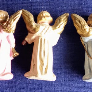 Puede incluir: Cinco peque&ntilde;as figuras de &aacute;ngeles con alas doradas y t&uacute;nicas en tonos pastel. Los &aacute;ngeles est&aacute;n dispuestos en fila sobre un fondo azul oscuro, resaltando sus delicados detalles.