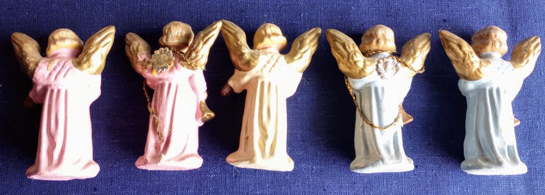Puede incluir: Cinco peque&ntilde;as figuras de &aacute;ngeles con alas doradas y t&uacute;nicas en tonos pastel. Los &aacute;ngeles est&aacute;n dispuestos en fila sobre un fondo azul oscuro, resaltando sus delicados detalles.