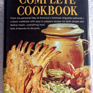 May include: A vintage cookbook cover featuring a roasted rack of lamb with white beans and herbs, a brown ceramic crock pot, and onions. The title of the book is "Amy Vanderbilt's Complete Cookbook".