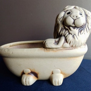 Può includere: Un vaso da fiori in ceramica a forma di vasca da bagno con una statuetta di leone seduta al suo interno. Il vaso è bianco con accenti marroni e il leone è bianco con la pelliccia marrone.