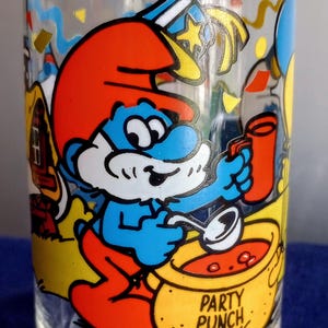 Puede incluir: Vaso transparente con una ilustración de Papá Pitufo de Los Pitufos. Papá Pitufo lleva un gorro rojo y está removiendo una olla de "Party Punch". El vaso tiene un diseño festivo con globos y confeti.