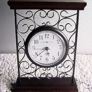 May include: Black clock with a white face and black Roman numeral hour markers. The clock is set in a black metal frame with a decorative scroll design. The clock is sitting on a wooden base.