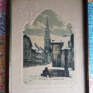 Op de afbeelding: Een ingelijste prent met een besneeuwd stadsplein met een hoge kerktoren, een stenen fontein en omliggende gebouwen. De rand is in reliëf versierd met sterren, hulst en de tekst MERRY XMAS in een gotische booglijst met een donkere houten rand.