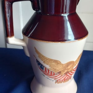 May include: A vintage ceramic pitcher with a brown top and handle, and a white body. The body features a patriotic design of an eagle with the American flag and the text "The Spirit of". The pitcher has a wide base and a spout.