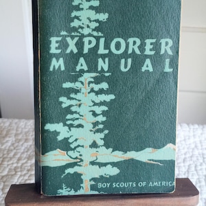 Op de afbeelding: Vintage Explorer Manual boek met een teal cover. De cover heeft een lichtblauwe illustratie van een dennenboom en de woorden "EXPLORER MANUAL" in grote, teal letters. De onderkant van het boek luidt "BOY SCOUTS OF AMERICA".