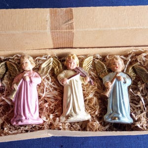 Puede incluir: Un conjunto de cinco figuras de &aacute;ngeles vintage en una caja de cart&oacute;n. Los &aacute;ngeles visten t&uacute;nicas rosas, crema y azules, con alas doradas y cabello rubio. Cada &aacute;ngel sostiene un instrumento musical, como un viol&iacute;n y una trompeta.
