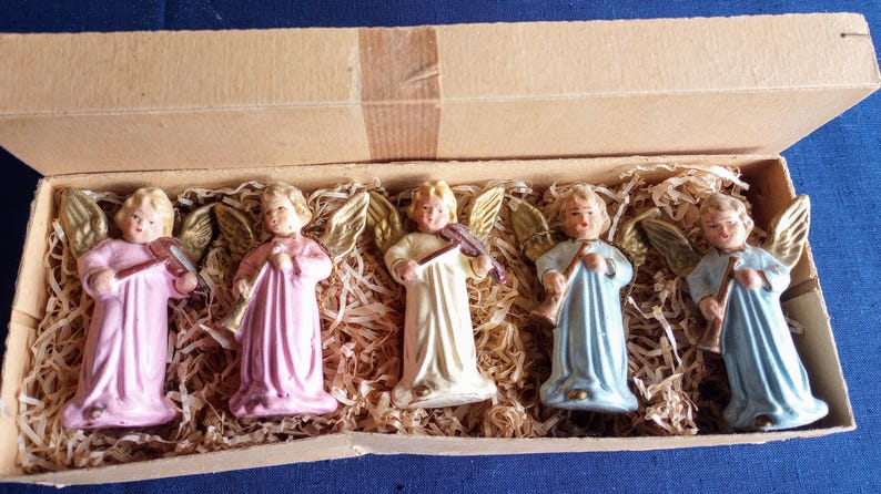 Puede incluir: Un conjunto de cinco figuras de &aacute;ngeles vintage en una caja de cart&oacute;n. Los &aacute;ngeles visten t&uacute;nicas rosas, crema y azules, con alas doradas y cabello rubio. Cada &aacute;ngel sostiene un instrumento musical, como un viol&iacute;n y una trompeta.