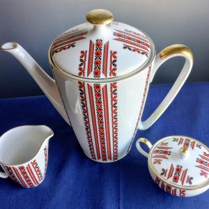 May include: A white porcelain tea set with red and black geometric patterns. The set includes a teapot, creamer, and sugar bowl, all with gold accents. The teapot has a lid and a long spout.