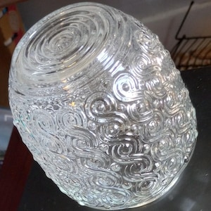 Puede incluir: Un jarrón de vidrio transparente con una superficie texturizada. El jarrón tiene una forma redondeada y está decorado con un patrón de remolino. La parte superior del jarrón tiene un diseño en espiral.