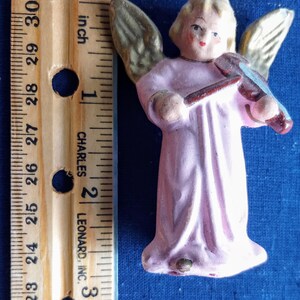 Puede incluir: Peque&ntilde;a figura de &aacute;ngel pintada a mano con una t&uacute;nica rosa claro, tocando el viol&iacute;n. El &aacute;ngel tiene alas doradas y una cabeza rubia. La figura mide aproximadamente 7 cm de altura, medida con una regla de madera.