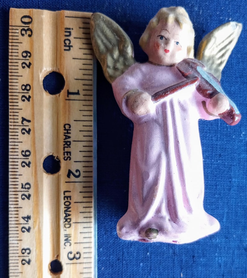 Puede incluir: Peque&ntilde;a figura de &aacute;ngel pintada a mano con una t&uacute;nica rosa claro, tocando el viol&iacute;n. El &aacute;ngel tiene alas doradas y una cabeza rubia. La figura mide aproximadamente 7 cm de altura, medida con una regla de madera.