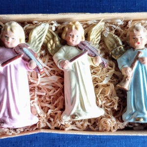 Puede incluir: Un conjunto de cinco figuras de &aacute;ngeles en una caja rectangular. Los &aacute;ngeles visten colores pastel: rosa, verde claro y azul. Cada &aacute;ngel tiene alas doradas y sostiene un instrumento musical. La caja est&aacute; llena de paja.