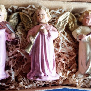 Puede incluir: Tres figuras de &aacute;ngeles de cer&aacute;mica con cabello rubio y alas doradas. Dos &aacute;ngeles visten t&uacute;nicas rosas y uno viste una t&uacute;nica color crema. Un &aacute;ngel toca la trompeta y dos tocan el viol&iacute;n. Las figuras se exhiben en una caja con relleno de papel.