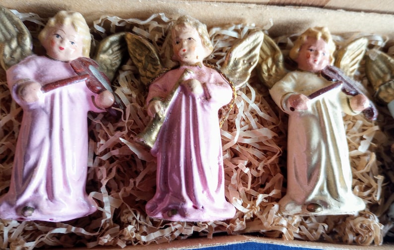 Puede incluir: Tres figuras de &aacute;ngeles de cer&aacute;mica con cabello rubio y alas doradas. Dos &aacute;ngeles visten t&uacute;nicas rosas y uno viste una t&uacute;nica color crema. Un &aacute;ngel toca la trompeta y dos tocan el viol&iacute;n. Las figuras se exhiben en una caja con relleno de papel.