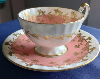 Puede incluir: Juego de taza y platillo de té vintage. La taza y el platillo son blancos con interiores rosas y detalles florales dorados. El borde y el asa de la taza también son dorados. El platillo tiene un centro rosa a juego y un diseño floral dorado.