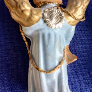 Puede incluir: Figura de &aacute;ngel vintage con alas doradas y una t&uacute;nica azul claro. El &aacute;ngel tiene un halo dorado y un detalle de flor plateada en la espalda. Un ribete dorado en forma de cuerda acent&uacute;a la figura.