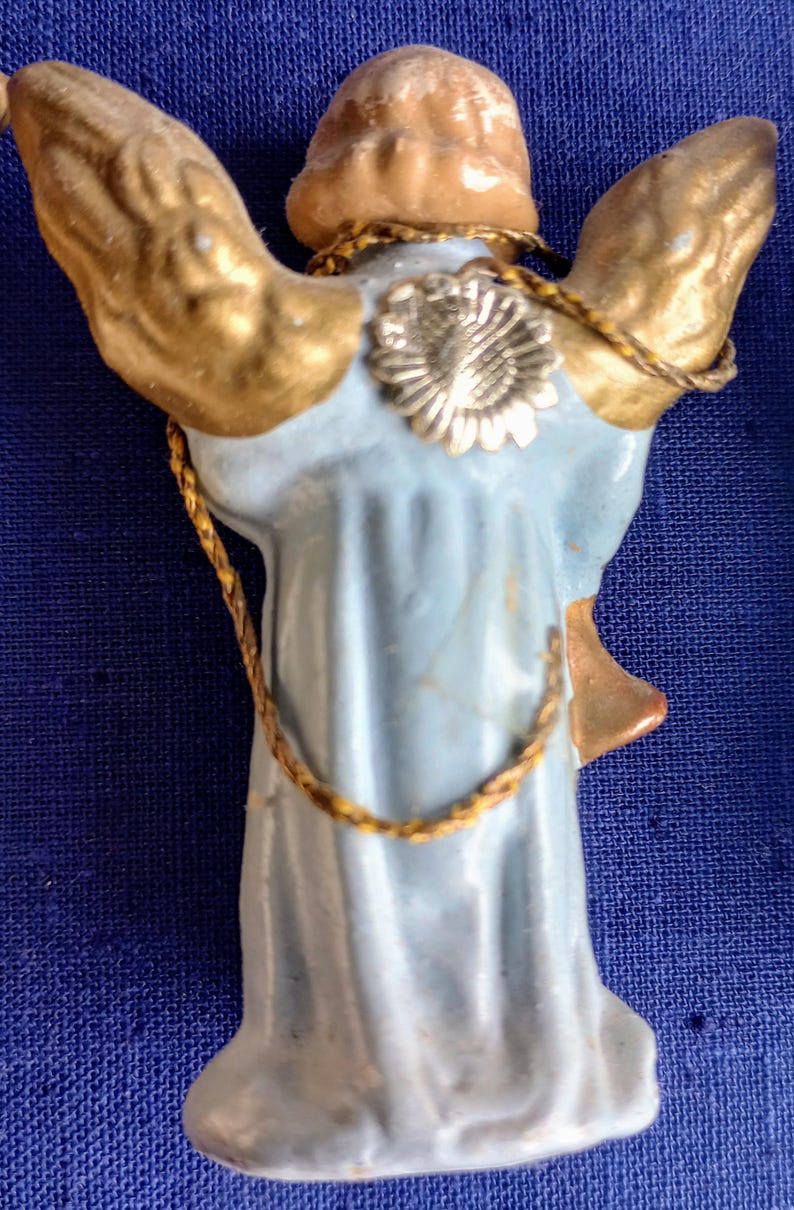 Puede incluir: Figura de &aacute;ngel vintage con alas doradas y una t&uacute;nica azul claro. El &aacute;ngel tiene un halo dorado y un detalle de flor plateada en la espalda. Un ribete dorado en forma de cuerda acent&uacute;a la figura.