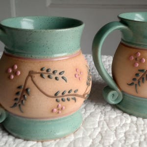 May include: Two ceramic mugs featuring a speckled mint green glaze on the rim and base, with a matte peach-colored center band. The center is decorated with embossed brown vine patterns and small pink flower accents, each with a curved handle.