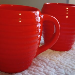 May include: Two bright red ceramic mugs with ribbed detailing. Each mug has a large, curved handle. The mugs are set on a white, textured surface. The mugs are a vibrant, solid color.