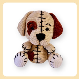 Puede incluir: Un juguete de peluche de perro tejido a crochet con pelaje marrón y blanco, orejas marrones y ojos negros. El perro está sentado con las piernas cruzadas y tiene una costura cosida en la espalda.