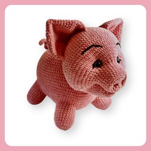 Puede incluir: Un juguete de cerdo de crochet rosa con ojos negros y una sonrisa cosida. El cerdo tiene una cola rizada y está de pie sobre sus cuatro patas.