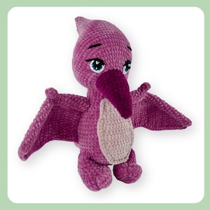 May include: A handmade, plush pterodactyl toy in shades of purple, with a lighter beige chest. The crochet dinosaur has large, blue eyes, a dark purple beak, and outstretched wings. The toy is designed to stand upright.
