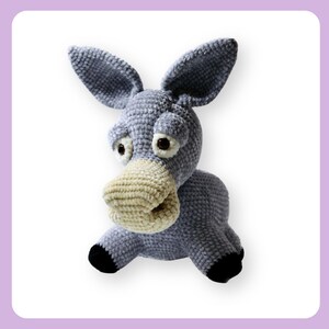 Puede incluir: Un juguete de peluche de burro de crochet gris con grandes ojos marrones y un hocico beige. El burro está sentado sobre un fondo blanco.