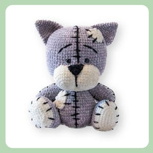 May include: A crocheted plush toy cat with a grey and white colour scheme. The cat has a stitched-on patch on its head and a stitched-on patch on its arm. The cat is sitting with its legs crossed.
