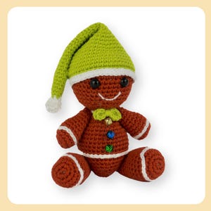 Puede incluir: Un juguete de jengibre de crochet que lleva un gorro de crochet verde y blanco. El juguete tiene un cuerpo marrón con ribete blanco, una pajarita verde y botones de colores.