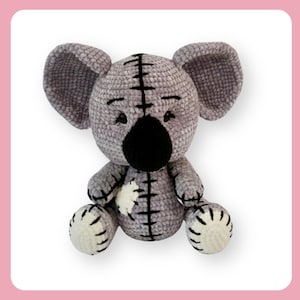 Puede incluir: Un juguete de peluche de koala de crochet gris con ojos y nariz negros, sentado con las piernas cruzadas. El juguete tiene detalles blancos en las orejas y los pies.