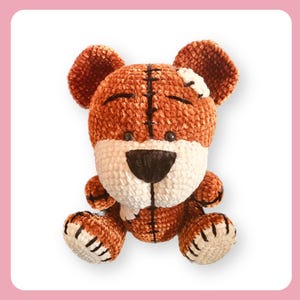 Op de afbeelding: Een bruine en witte gehaakte pluche beer met een gestikte patch op zijn hoofd. De beer heeft zwarte ogen en een zwarte neus.