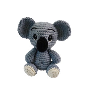 Puede incluir: Un juguete de peluche de koala de crochet gris con ojos negros, nariz negra y pies blancos. El koala está sentado con los brazos cruzados.