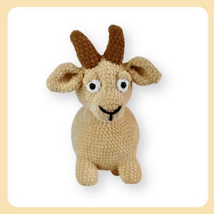 Puede incluir: Un juguete de peluche de cabra tejido a crochet con cuernos marrones, ojos blancos y una nariz negra. La cabra está sentada sobre un fondo blanco.