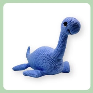 May include: A blue crocheted Nessie toy, featuring a long neck, small flippers, and a smiling face. The handmade plush toy is a light shade of blue and rests on a white background.