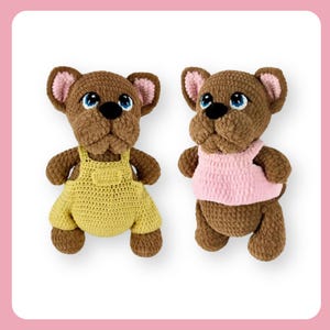 Op de afbeelding: Twee gehaakte bruine pluche hondenspeelgoedjes die verschillende outfits dragen. Eén hond draagt een gele overall en de andere een roze tanktop.