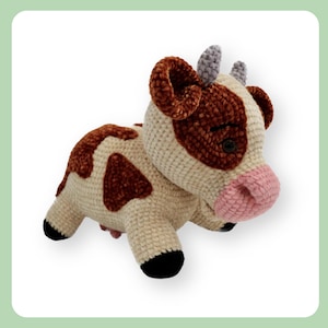 Puede incluir: Un peluche de vaca hecho a mano con ganchillo. La vaca tiene un cuerpo color crema con manchas y detalles marrones, incluyendo las orejas y una mancha alrededor de los ojos. Tiene un hocico rosa, ojos negros y pezuñas negras. Los cuernos son grises.