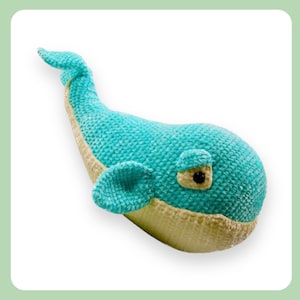 Puede incluir: Un juguete de peluche de ballena de crochet con un cuerpo azul claro y una barriga de color crema. La ballena tiene un solo ojo negro y una pequeña aleta azul claro.