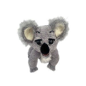 Puede incluir: Un juguete de peluche de koala gris tejido a crochet con ojos negros, nariz negra y pecho blanco y esponjoso. El koala está de pie sobre sus cuatro patas con la cabeza ligeramente inclinada hacia un lado.