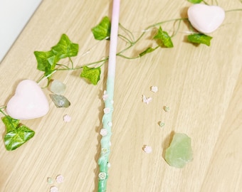 May include: A pastel-colored wand with a gradient of pink to green, adorned with small pink flowers. The wand is decorated with small, light pink flowers. The image also includes a heart-shaped object, green leaves, and small crystals.