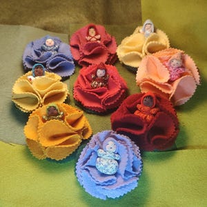 May include: A collection of eight small dolls, each nestled within a ruffled felt flower. The dolls feature varied skin tones and patterned fabric clothing, while the felt flowers come in shades of blue, red, yellow, orange, and purple.