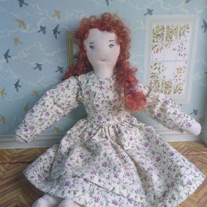 May include: A fabric doll with light skin, blue embroidered eyes, and long curly red hair. The doll wears a long-sleeved dress featuring a cream-colored background with a repeating pattern of small green vines and tiny purple flowers.