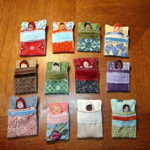 Puede incluir: Once muñecos de tela pequeños metidos en sacos de dormir en miniatura individuales. Cada muñeco tiene un color de pelo y una expresión facial únicos, mientras que los sacos de dormir presentan varios patrones, incluyendo flores, cachemir, geométricos y colores sólidos.